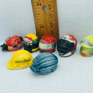 Può includere: Una collezione di sette caschi in miniatura in ceramica di vari colori e design, tra cui un casco rosso e bianco con una torcia frontale, un casco nero e verde con la scritta "Honda", un casco rosso con la scritta "Trofeo", un casco blu e rosso con una maschera facciale, un casco giallo con un design da pompiere, un casco da cantiere giallo con la scritta "Novodimet" e un casco blu con un design a righe.