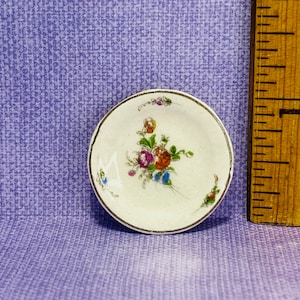May include: A small white porcelain plate with a floral design. The plate has a gold rim and features a cluster of pink, red, and blue flowers with green leaves.