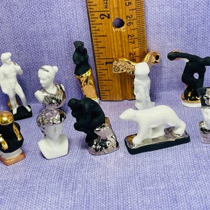 May include: A collection of miniature ceramic figurines, including busts, a polar bear, and a male figure. The figurines are painted in white, black, gold, and silver, and are arranged on a purple surface. A wooden ruler is also present.
