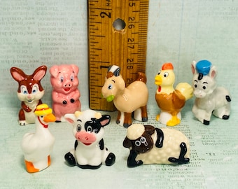 May include: A collection of eight small ceramic figurines of farm animals. The figurines are painted in various colors, including brown, pink, white, black, and gray. The animals include a rabbit, pig, goat, chicken, donkey, goose, cow, and sheep.