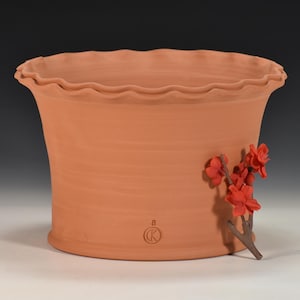 Puede incluir: Una maceta de cerámica color terracota con un borde ondulado y una forma acampanada. Una pequeña rama con flores rojas está adherida al lateral. La maceta tiene un logotipo circular y el número 8.