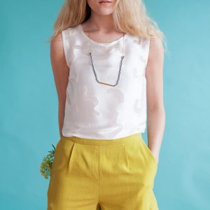 Puede incluir: Una mujer con un top blanco sin mangas, un collar dorado y negro y pantalones cortos amarillos. Est&aacute; de pie frente a un fondo azul.