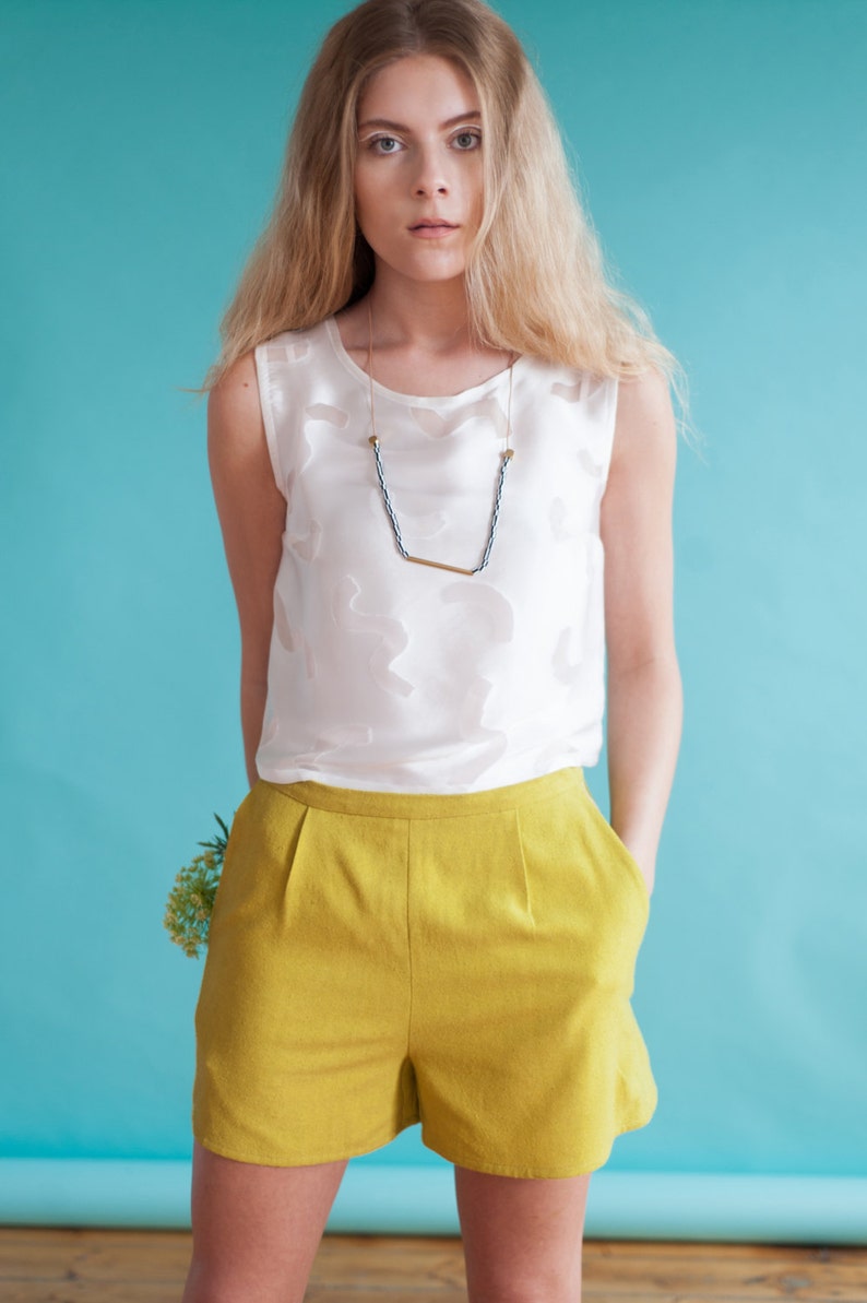 Puede incluir: Una mujer con un top blanco sin mangas, un collar dorado y negro y pantalones cortos amarillos. Est&aacute; de pie frente a un fondo azul.