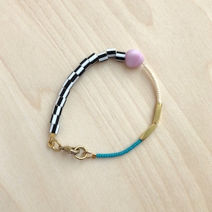 May include: A black and white chequered bracelet with a pink bead and gold accents. The bracelet has a turquoise blue section and a gold clasp.
