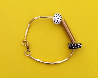 May include: A gold bracelet with black and white polka dot beads. The bracelet has a unique design with a long, straight piece of metal connecting two of the beads.