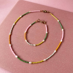 May include: A set of two necklaces with a multi-coloured beaded design. The necklace is made of small beads in shades of green, yellow, pink, and light blue. The necklace is accented with small white pearl beads. The set includes a shorter necklace and a longer necklace.