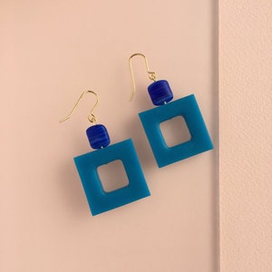 Op de afbeelding: Een paar blauwe vierkante oorbellen met een kleinere vierkante uitsparing in het midden. Elke oorbel heeft bovenaan een blauwe en witte gestreepte kraal.