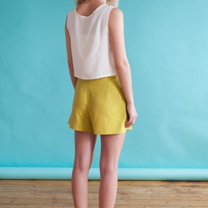 Puede incluir: Una mujer con un top blanco sin mangas y pantalones cortos amarillos. Est&aacute; de pie frente a un fondo azul y mira hacia la derecha.