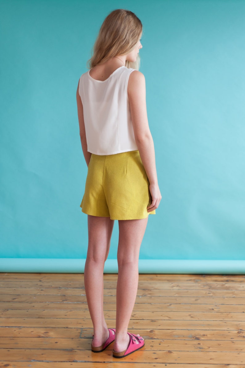 Puede incluir: Una mujer con un top blanco sin mangas y pantalones cortos amarillos. Est&aacute; de pie frente a un fondo azul y mira hacia la derecha.