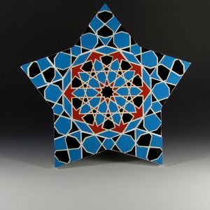 May include: A star-shaped decorative piece with a geometric pattern. The design features blue, black, red, and white shapes arranged in a symmetrical pattern. The central design is a complex starburst pattern. The piece is set against a gradient background.