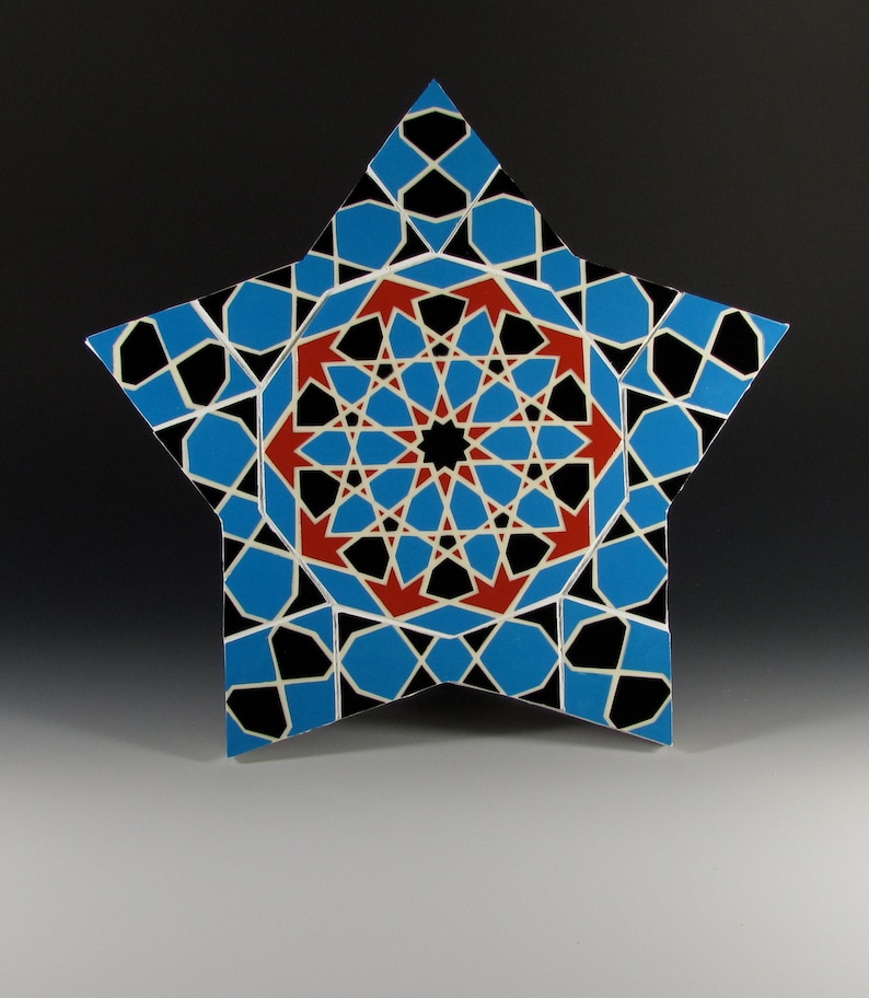 May include: A star-shaped decorative piece with a geometric pattern. The design features blue, black, red, and white shapes arranged in a symmetrical pattern. The central design is a complex starburst pattern. The piece is set against a gradient background.