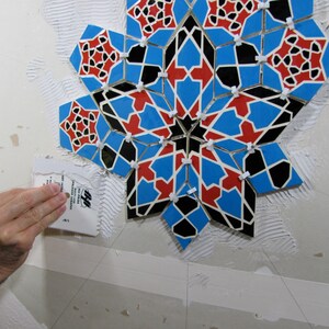 May include: A mosaic tile installation in progress. The design features a geometric star pattern with blue, red, and black tiles. A hand uses a trowel to apply adhesive to the wall, preparing for the tile placement.