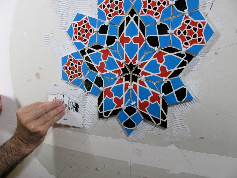 May include: A mosaic tile installation in progress. The design features a geometric star pattern with blue, red, and black tiles. A hand uses a trowel to apply adhesive to the wall, preparing for the tile placement.