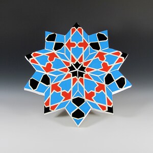 May include: A geometric star-shaped decorative object. The design features a symmetrical pattern with blue, red, black, and white segments. The object is likely made of wood or a similar material, with a painted finish. The star has a complex, multi-pointed shape.