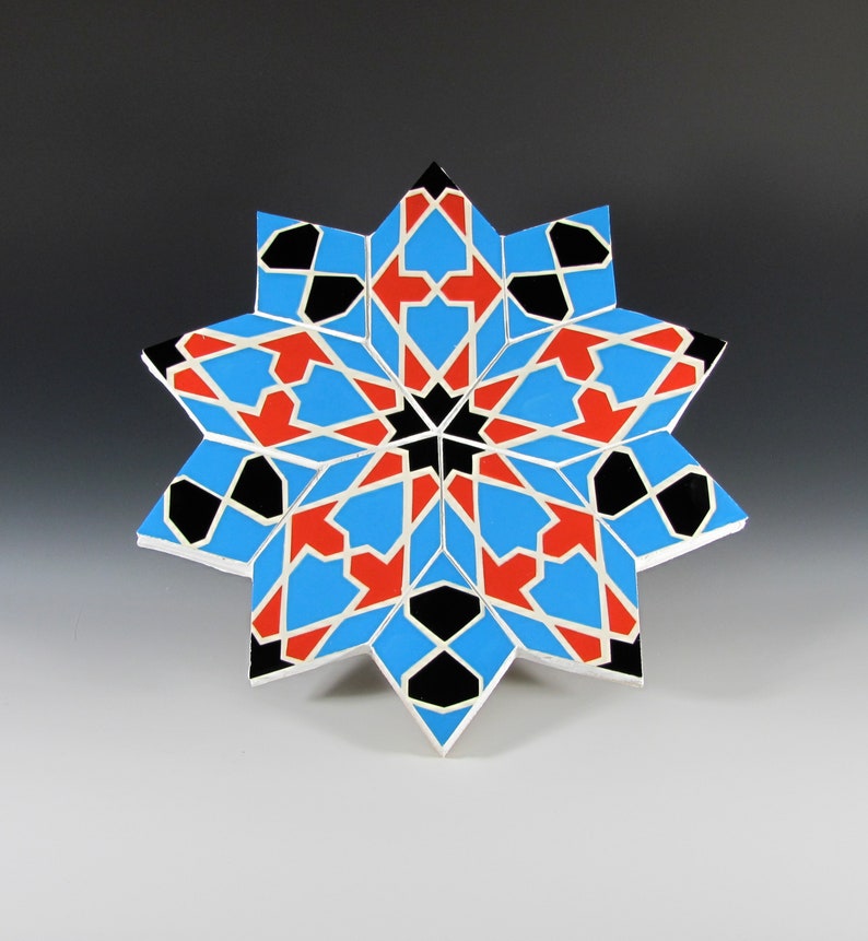 May include: A geometric star-shaped decorative object. The design features a symmetrical pattern with blue, red, black, and white segments. The object is likely made of wood or a similar material, with a painted finish. The star has a complex, multi-pointed shape.