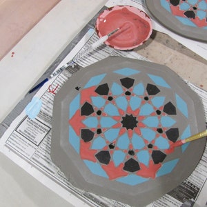 May include: A ceramic plate with a geometric design in progress. The plate features a gray base with a pattern of blue, red, and black shapes. A small bowl of pink paint and a paintbrush are nearby. The plate is on a newspaper.