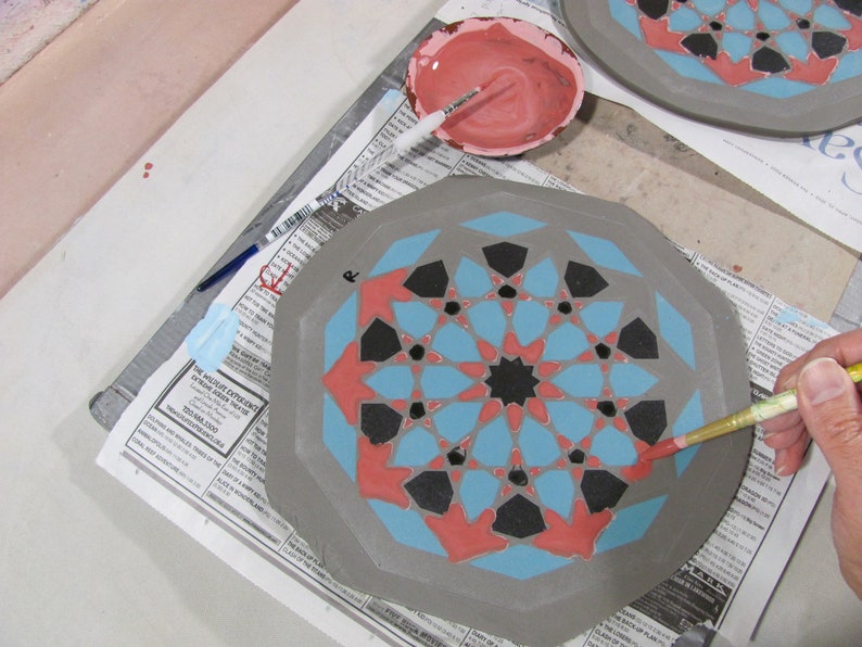 May include: A ceramic plate with a geometric design in progress. The plate features a gray base with a pattern of blue, red, and black shapes. A small bowl of pink paint and a paintbrush are nearby. The plate is on a newspaper.