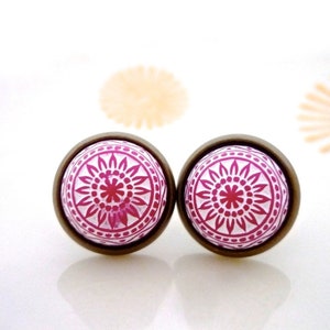 May include: A pair of round stud earrings. Each earring features a white background with a magenta floral design. The design is encased in a bronze-colored metal setting. The earrings are displayed on a white surface.