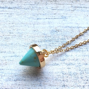 May include: A gold-colored chain necklace with a turquoise-colored, cone-shaped pendant. The pendant is encased in a gold-colored metal setting. The necklace is displayed on a light-colored, textured surface.