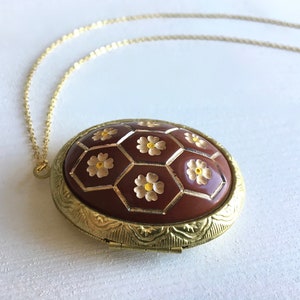 May include: A gold chain necklace with a round locket pendant. The locket is brown with a gold rim and features a design of white flowers on a honeycomb pattern.