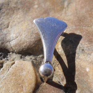 May include: A silver-colored metal pendant shaped like a fan with a rounded bottom. The pendant is on a textured surface.