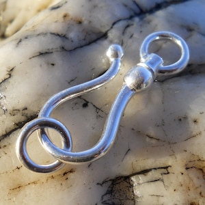 May include: Silver-toned metal clasp with a round ring and a hook. The clasp is on a white, textured surface.