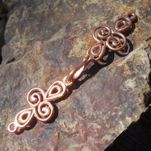 May include: A copper-colored metal clasp with a decorative design of interlocking swirls. The clasp is designed to connect two pieces of jewelry or fabric.