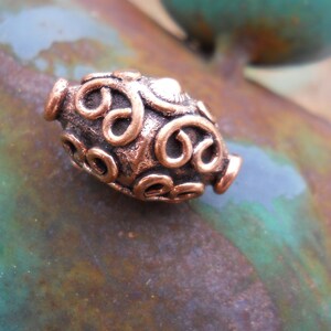 May include: A copper-colored bead with a swirling, intricate design. The bead is oval-shaped and has a smooth, polished finish.