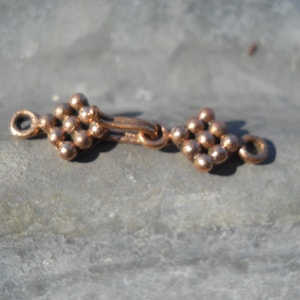 May include: A copper-colored metal clasp with two square-shaped pieces connected by a loop. Each square is made of small, round beads.