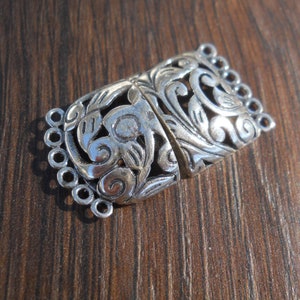 May include: Silver-toned metal clasp with an ornate floral design. The clasp has five loops on one side for attaching jewelry.