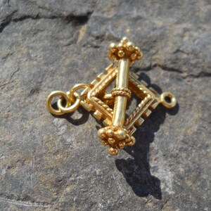 May include: A gold-colored pendant with a central vertical bar and a diamond-shaped frame. The pendant features intricate detailing, including a twisted rope design and small, spherical accents. Two loops are attached for hanging.