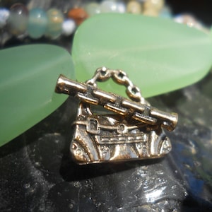 May include: A gold-toned charm in the shape of a handbag with a detailed design. The charm has a chain and a small, detailed, gold-toned  handle. The charm is sitting on a green surface.