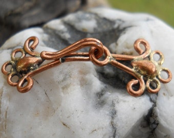 May include: A copper-colored metal clasp with a decorative design. The clasp is made of two pieces that are joined together with a hook.