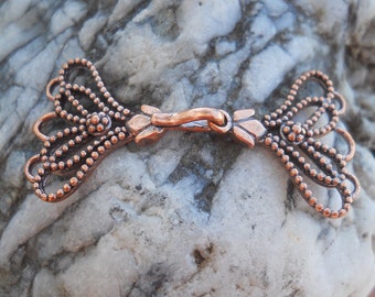 May include: A copper-colored metal clasp with a decorative design. The clasp has a rounded center section and two ornate, fan-shaped ends.