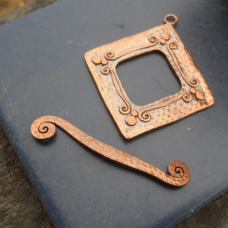 Op de afbeelding: Twee koperen sieraden met een gehamerde textuur en een swirl-ontwerp. Een stuk is een vierkant frame met een gat aan de bovenkant om op te hangen. Het andere stuk is een gebogen vorm met een swirl aan elk uiteinde.
