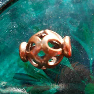 May include: A close-up of a small, intricate, copper-colored metal ornament with a geometric design. The ornament is shaped like a sphere with a circular opening on one side. The ornament is resting on a green surface.