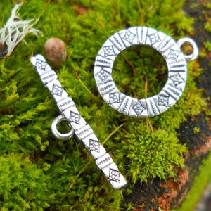 May include: Silver tone toggle clasp with a geometric design. The clasp has a round ring and a bar with a loop.