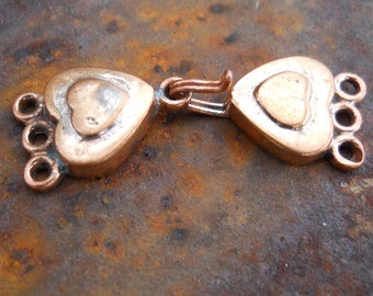 May include: A copper-toned heart-shaped clasp with a decorative design. The clasp has a loop on each side and a series of small loops on one side.