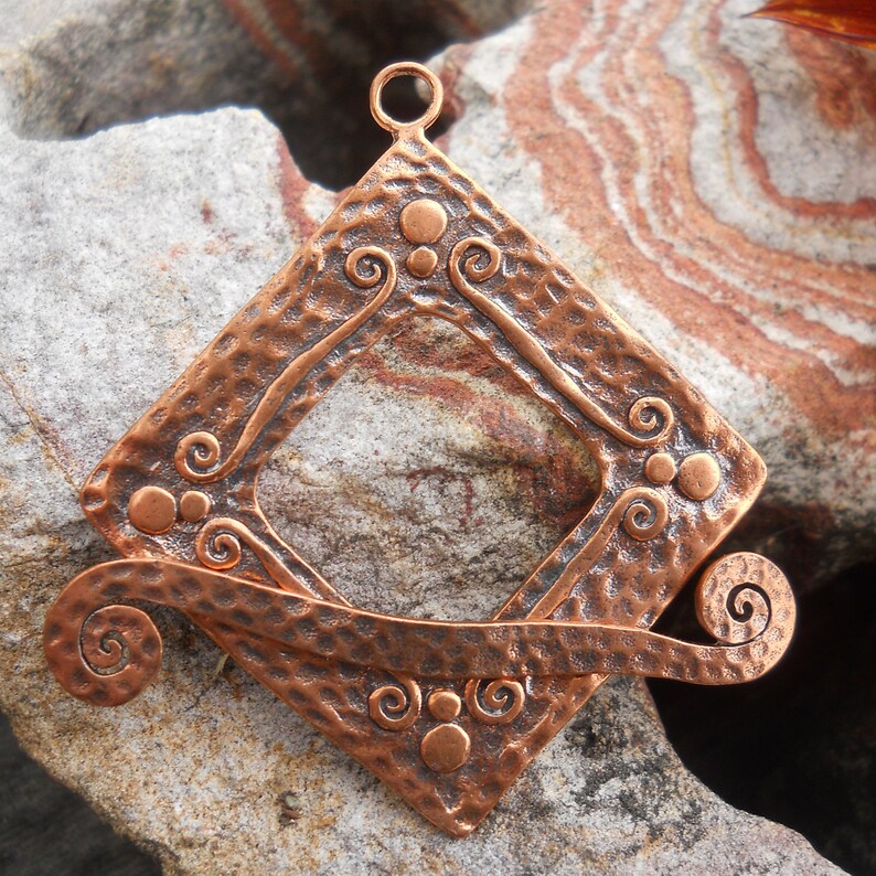 Op de afbeelding: Een koperen hanger met een gehamerde afwerking en een vierkant frame met een uitsparing in het midden. Het frame is versierd met spiralen en stippen. De hanger heeft een lus aan de bovenkant om op te hangen.