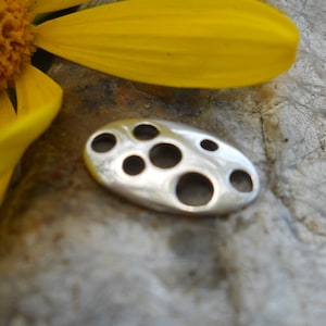 May include: A silver-colored, oval-shaped pendant with seven circular cutouts. The pendant is smooth and reflective, resting on a textured, gray surface. A yellow flower petal is visible in the background.