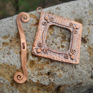 Op de afbeelding: Twee koperen metalen stukken met een gehamerde textuur. Een stuk is een vierkant frame met een decoratief swirl-ontwerp. Het andere stuk is een lang, dun stuk met een swirl aan elk uiteinde.