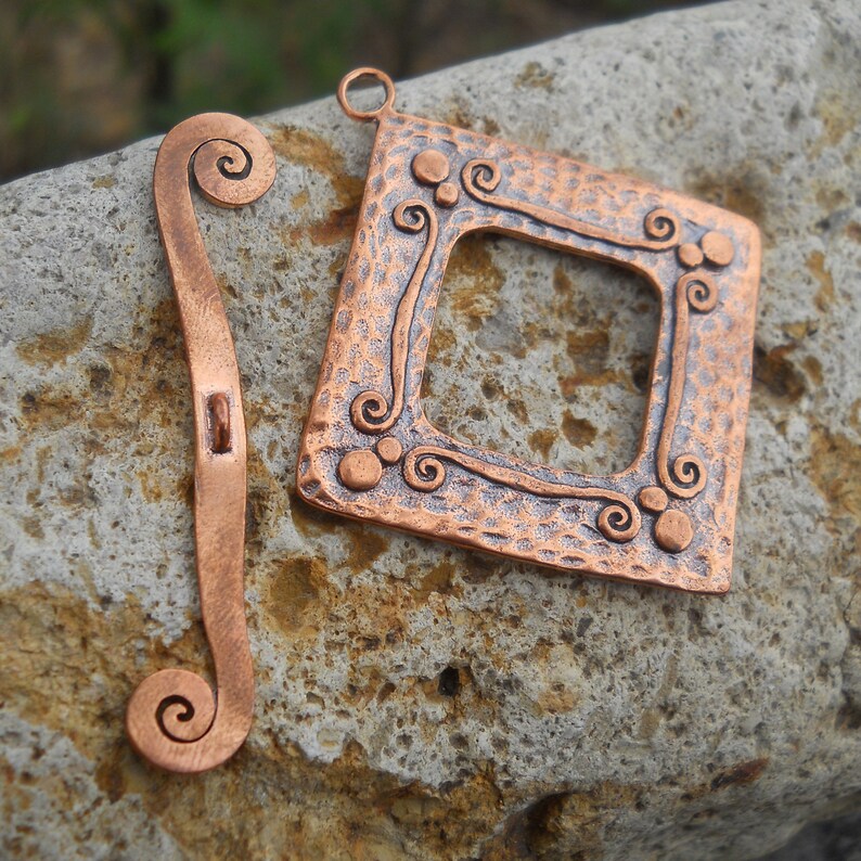 Op de afbeelding: Twee koperen metalen stukken met een gehamerde textuur. Een stuk is een vierkant frame met een decoratief swirl-ontwerp. Het andere stuk is een lang, dun stuk met een swirl aan elk uiteinde.