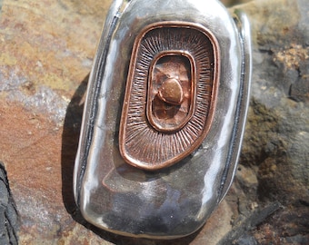 May include: A silver-toned pendant with a copper-colored, layered, oval-shaped design in the center. The pendant has a smooth, polished surface and is set against a textured, brown background.