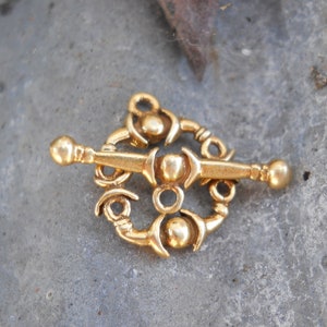 May include: A gold-colored metal pendant with a circular design featuring intricate details and three round knobs. The pendant has a long, thin bar with rounded ends.