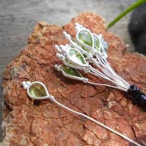 May include: A set of silver hair pins with green gemstone accents. The pins are arranged in a fan shape and are displayed on a brown, textured surface.