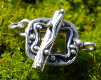 May include: Silver-toned metal clasp with a decorative design featuring a stylized branch and a square frame. The clasp has a textured surface and is designed for jewelry making.