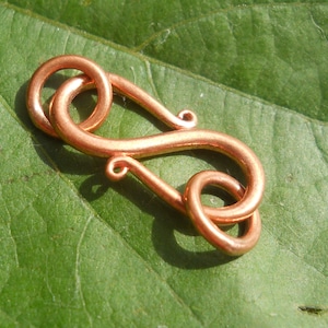 May include: A copper-colored metal clasp with a decorative, intricate design. The clasp is shaped like a figure eight with a small loop at the top.