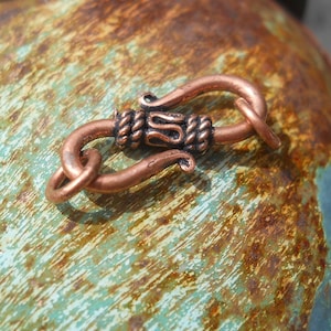 May include: A handcrafted copper hook clasp, featuring a decorative braided center and curved ends. The clasp is designed for jewelry making or other craft projects. The copper has a warm, reddish-brown hue, and the clasp is shown against a textured, turquoise and brown background.