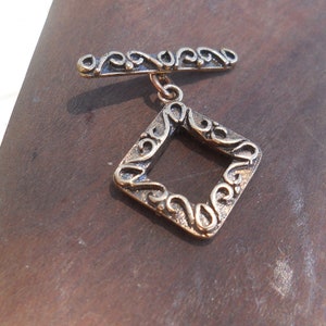 May include: A gold-toned metal clasp with a square frame design. The frame is decorated with intricate swirls and has a small loop at the top.