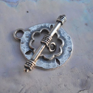 Op de afbeelding: Zilverkleurige metalen toggle-sluiting met een decoratief, gegolfd cirkelontwerp. De sluiting heeft een gestructureerd oppervlak en een kleine lus aan de zijkant.